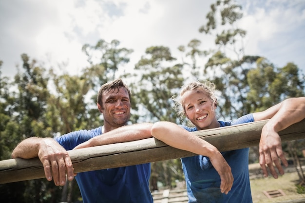 Sourire homme et femme s'appuyant sur un obstacle pendant la course d'obstacles dans le camp d'entraînement