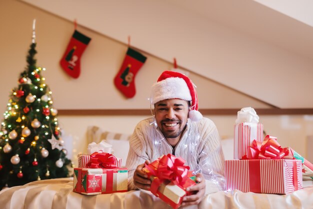 Sourire homme métis avec le chapeau du père Noël sur la tête posant sur le lit avec un cadeau dans ses mains.