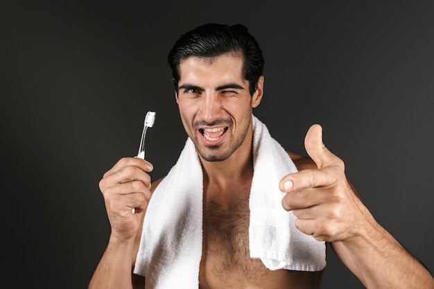 Sourire homme torse nu avec une serviette sur ses épaules debout isolé, tenant une brosse à dents