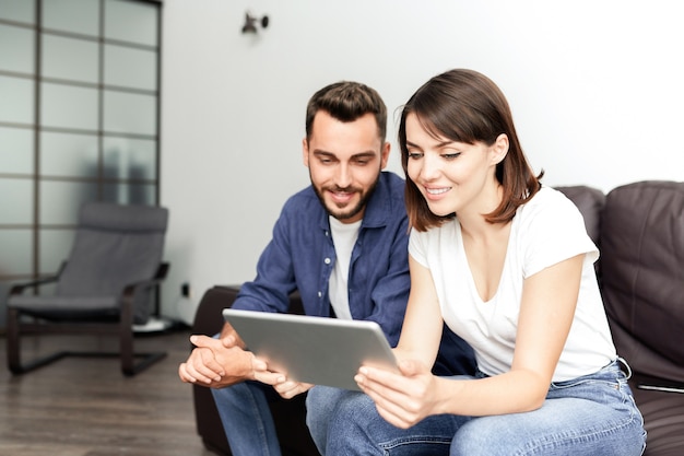 Sourire jeune couple assis sur un canapé confortable dans le salon et à l'aide de tablette pour naviguer sur Internet