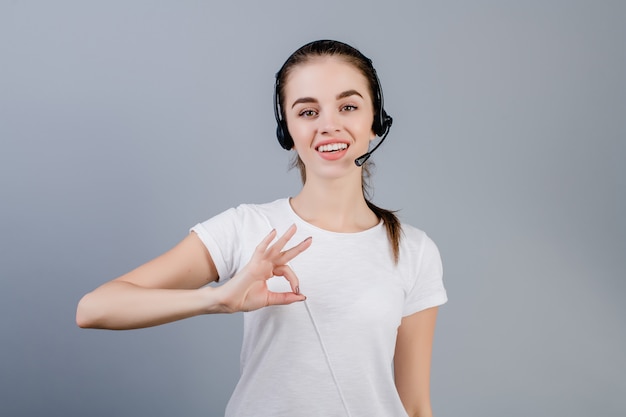 Sourire jeune femme de centre d'appel dispatcher portant casque répondant à la clientèle montrant le geste correct isolé sur gris