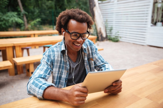 Sourire jeune homme à lunettes assis et à l'aide de tablette dans un café en plein air