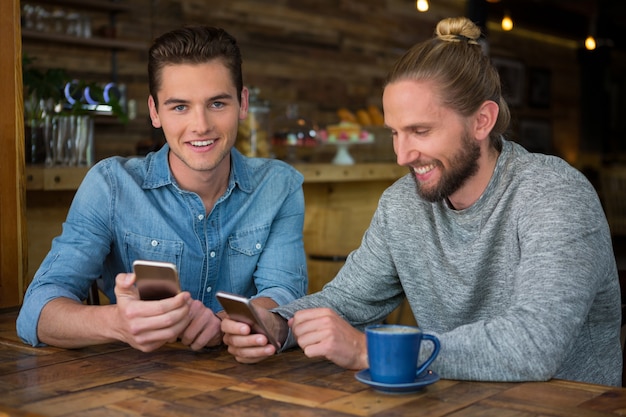 Sourire de jeunes hipsters masculins à l'aide de téléphones intelligents à table dans un café