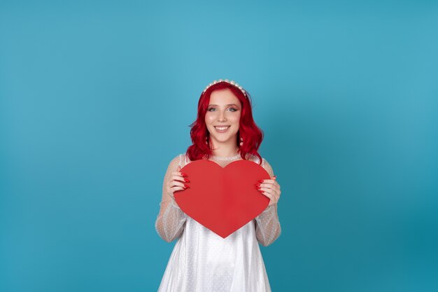 Sourire joyeuse jeune femme dans une robe blanche et aux cheveux rouges tient un grand coeur de papier rouge