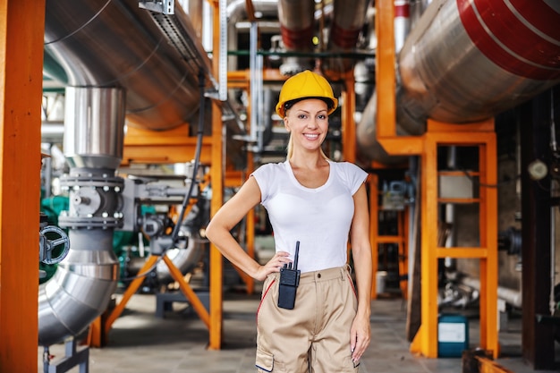 Sourire joyeuse travailleuse indépendante réussie en costume de travail et avec casque de protection sur la tête debout dans une installation de chauffage et regardant la caméra. Femme faisant le concept de travail masculin.