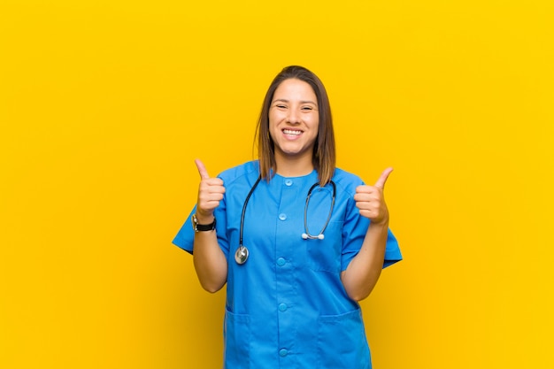 Sourire joyeusement et avoir l'air heureux, se sentir insouciant et positif avec les deux pouces levés, isolés contre un mur jaune