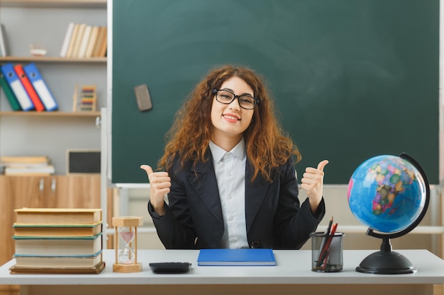 sourire montrant les pouces vers le haut jeune enseignante portant des lunettes assis au bureau avec des outils scolaires en classe