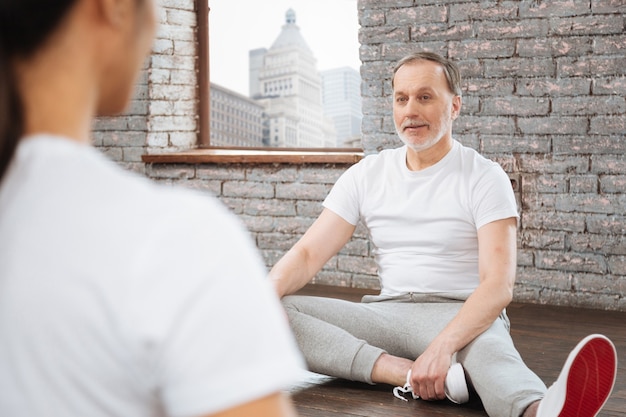 Sourire de personne de sexe masculin aux cheveux gris portant un T-shirt blanc et un pantalon gris mettant ses mains sur des baskets tout en regardant sur le formateur