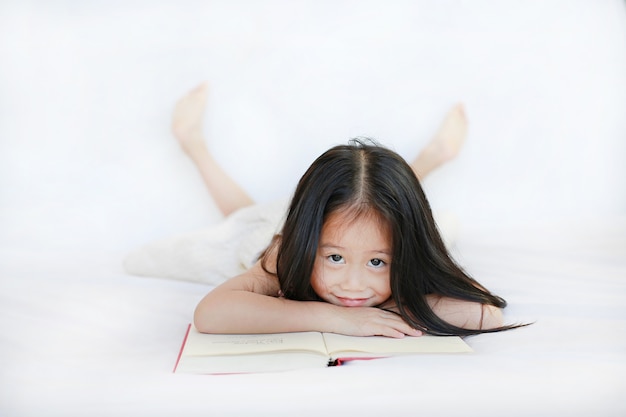 Sourire de petite fille asiatique avec livre à couverture rigide allongé sur le lit et regarder la caméra sur fond blanc.