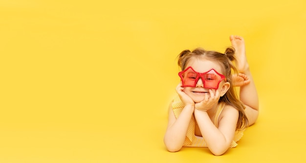 Sourire petite fille enfant vêtue d'un maillot de bain et lunettes de soleil drôle rouge allongé sur le mur jaune