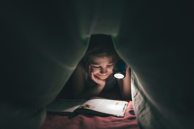 Sourire petite fille est lu un livre sous une couverture avec une lampe de poche dans une pièce sombre la nuit, au lit