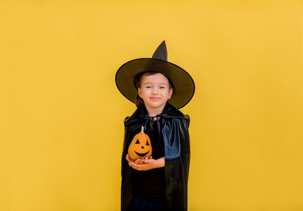 Sourire de petite sorcière avec un chapeau et Cape détient une citrouille et sourit sur un jaune isolé