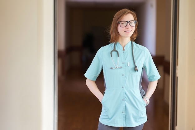 Sourire portrait de femme médecin rousse blanche avec stéthoscope sur le cou et les mains dans les poches