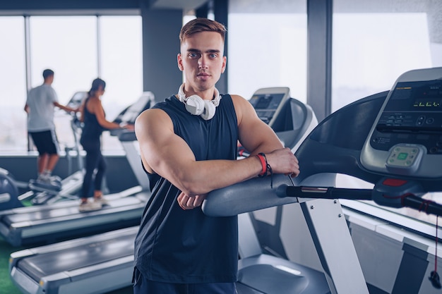 Sourire positif confiant instructeur personnel masculin avec bras croisés bras près de tapis roulant au gymnase dans la salle de fitness.