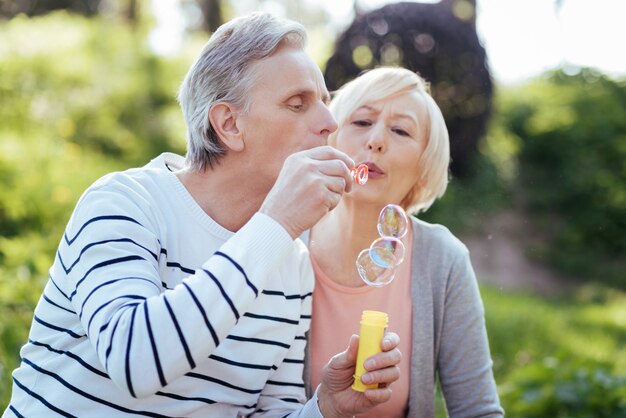 Sourire positif couple de personnes âgées s'embrassant et faisant des bulles de savon tout en profitant du temps libre dans le parc et en exprimant la joie