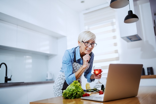 Sourire, séduisant, blond, femme aînée, dans, tablier, utilisation, ordinateur portable, pour, recette, et, boire vin