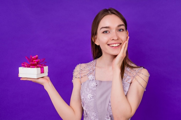 Sourire surpris belle femme mariée avec maquillage tenant une boîte-cadeau en studio sur fond violet. concept d'anniversaire et de fête de mariage