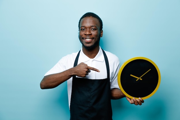 Sourire tenant et pointe à l'horloge murale jeune coiffeur afro-américain en uniforme isolé sur fond bleu