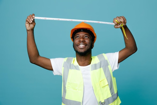 Sourire tenant ruban à mesurer jeune constructeur afro-américain en uniforme tenant isolé sur fond bleu