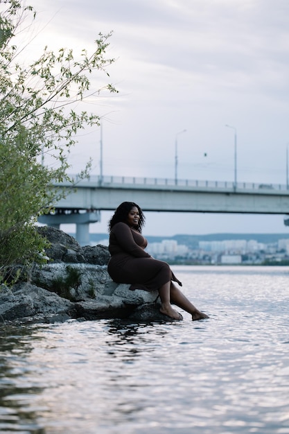 Sourire vertical serein pieds nus bouclés femme multiraciale afro-américaine dodue s'asseoir au bord de la rivière vacances tranquilles