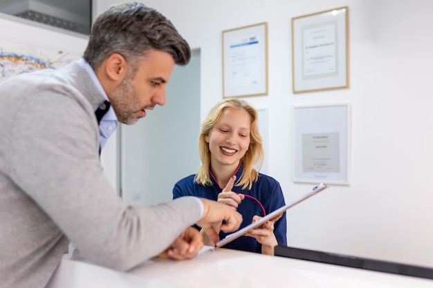Soutenir les patients Un homme souriant et son dentiste joyeux remplissent ensemble un formulaire à la réception