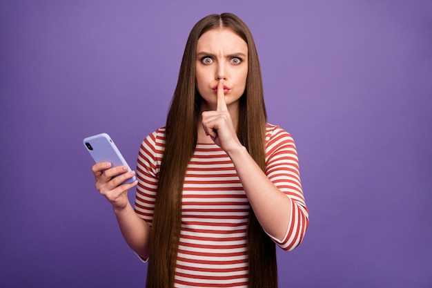 Soyez tranquille, ne partagez pas! Les jeunes filles sérieuses utilisent le smartphone ont des médias sociaux privés de communication montrent des lèvres d'index de signe sans voix porter cavalier rayé blanc mur de couleur violet