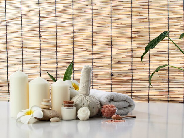 Spa Massage Concept, boule de compression aux herbes, crème, savon de fleur, bougie parfumée sur une table blanche, rideau de bambou