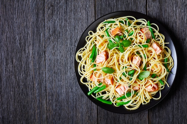 Spaghetti aux haricots verts et morceaux de saumon rôti, décorés de feuilles de basilic frais, servi sur une assiette noire sur une table en bois sombre, vue de dessus, mise à plat, espace copie