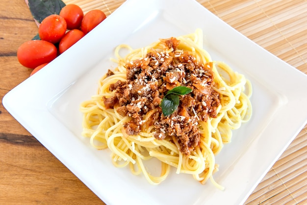 Spaghetti bolognaise servi sur une assiette blanche sur un fond en bois avec des tomates et du basilic.
