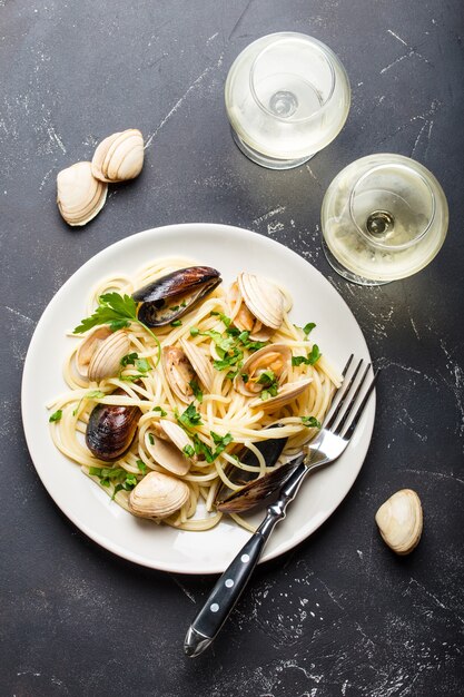 Spaghetti vongole, pâtes de fruits de mer italiennes aux palourdes et moules, en assiette avec des herbes et deux verres de vin blanc sur fond de pierre rustique.