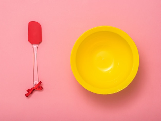 Spatule avec ruban rouge et bol jaune sur fond rose. Outils de cuisine sur fond festif. Mise à plat.