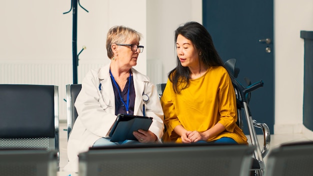 Spécialiste de la santé consultant un patient avec des béquilles, prenant des notes sur une tablette numérique pour faire des rapports médicaux. Diverses femmes âgées faisant un rendez-vous de consultation dans le hall de l'établissement.