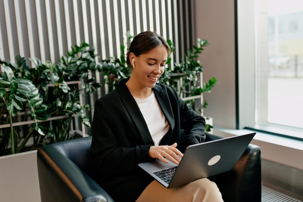 Spectaculaire femme adorable avec un magnifique sourire vêtu d'une veste sombre et d'un t-shirt blanc travaille avec un ordinateur portable dans un bureau moderne et élégant