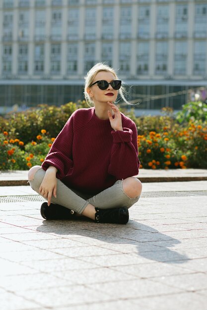 Splendide jeune femme portant un pull et des lunettes de soleil assis sur le trottoir