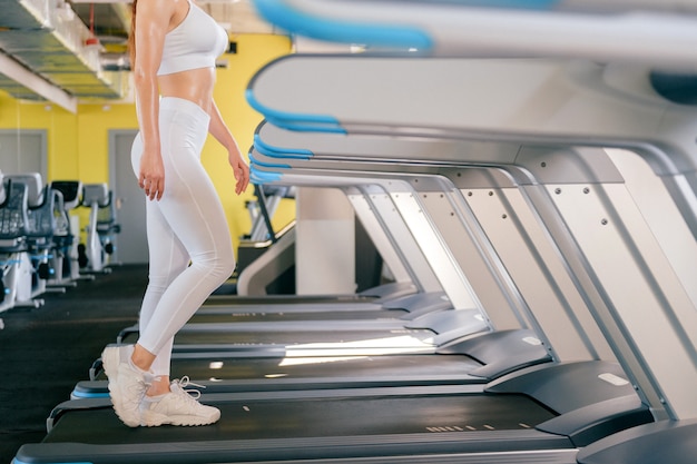 Sport femme qui court sur tapis roulant dans la salle de sport, garde la forme, brûle des calories sur la machine en cours d'exécution, portant des vêtements de sport blancs et des baskets.