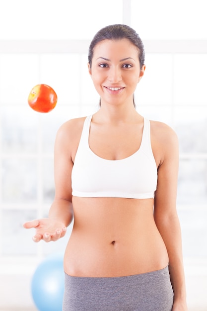 Sport et régime. Belle jeune femme en vêtements de sport jetant une pomme et souriant à la caméra
