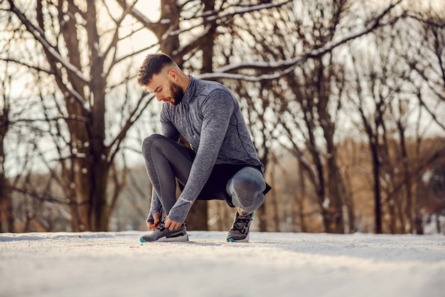 Sportif accroupi et attachant des lacets dans la nature au jour d'hiver enneigé. Vêtements de sport, fitness hiver, temps froid