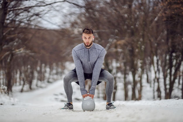 Sportif accroupi sur un chemin enneigé et soulevant kettlebell Fitness en plein air fitness hiver musculation