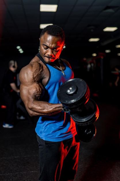 Sportif afro-américain professionnel posant devant la caméra en vêtements de sport et haltères à la main Fond de gym noir Corps parfait musclé