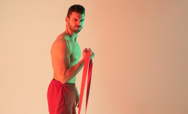 Sportif européen fort faisant de l'exercice avec une bande de résistance. Jeune bel homme barbu avec torse sportif nu. Isolé sur fond beige avec feu vert. Tournage en studio. Espace de copie