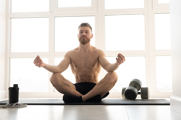 Sportif européen pratiquant le yoga et méditant sur un tapis de fitness. Jeune homme barbu au torse nu. Concept d'activité sportive à la maison. Intérieur d'un appartement spacieux et moderne. Journée ensoleillée