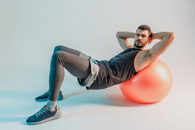 Le sportif fait des pompes sur un ballon de fitness. Un jeune homme européen barbu sérieux porte un uniforme de sport et regarde la caméra. Isolé sur fond gris avec une lumière turquoise. Tournage en studio. Espace de copie