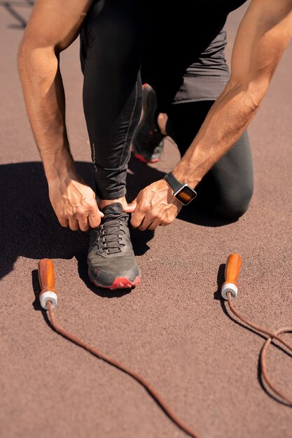 Sportif musclé en vêtements de sport noirs attachant les lacets sur sneaker avant de faire de l'exercice avec corde à sauter