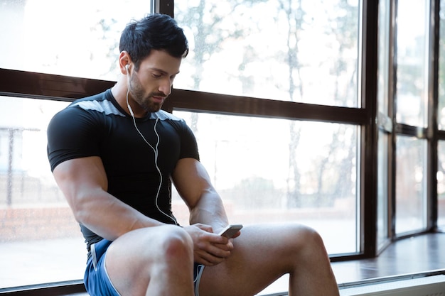 Sportif portant un short bleu et un t-shirt noir écoutant de la musique, à l'aide de son téléphone portable assis sur le rebord de la fenêtre