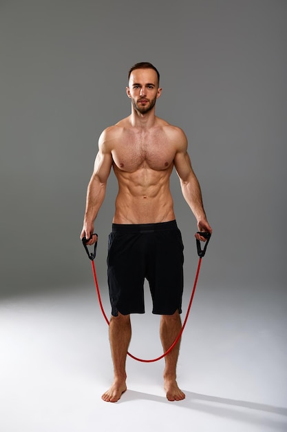 Un sportif seins nus effectue des exercices de fitness avec un élastique dans un studio sur fond gris
