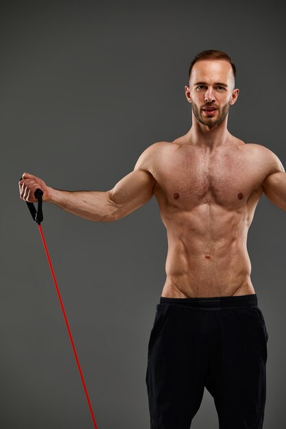 Un sportif seins nus effectue des exercices de fitness avec un élastique dans un studio sur fond gris