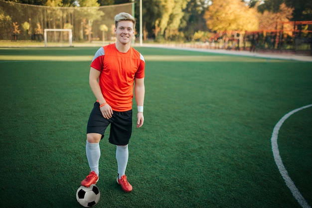 Un sportif sympa et bien bâti se tient debout et pose devant la caméra. Il tient un pied sur le ballon. Guy a l'air confiant. Il se tient sur la pelouse.