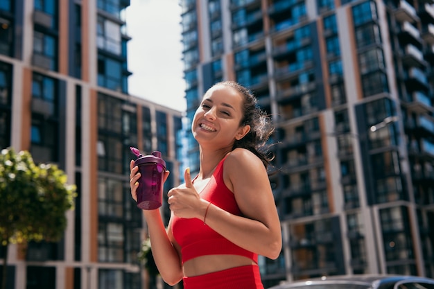 Sportive adorable jeune femme souriante aux cheveux noirs portant un soutien-gorge rouge tenant une bouteille de sport et le pouce en l'air