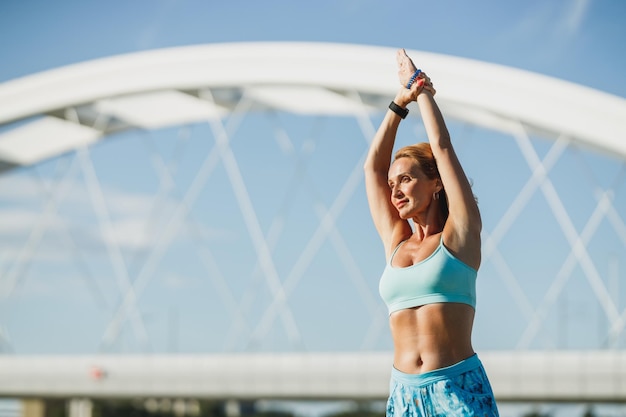 Sportive d'âge moyen faisant des exercices d'étirement tout en s'échauffant pour l'entraînement sportif près du pont de la rivière.