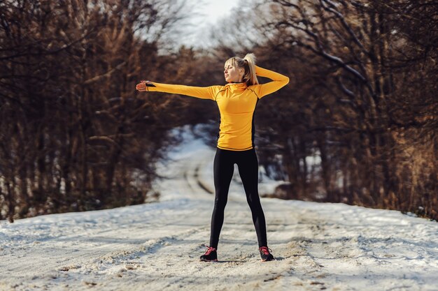 Sportive en forme debout sur un chemin enneigé dans la nature et faire des exercices d'échauffement lors d'une journée d'hiver ensoleillée.